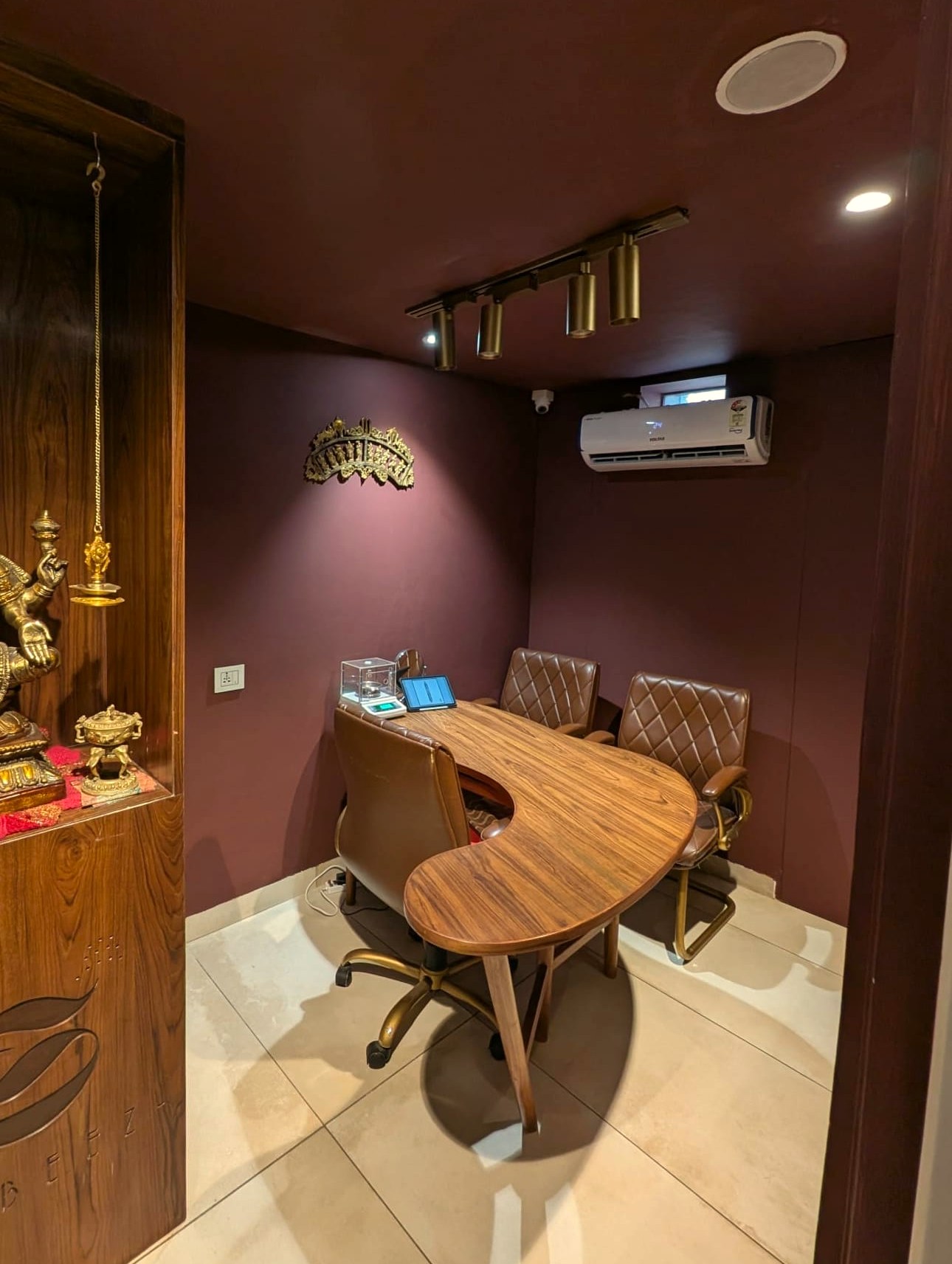 The Subeez viewing room: plum walls, a kidney-shaped teak table with a jeweller’s scale, leather chairs, and the brass Lakshmi in her wooden alcove