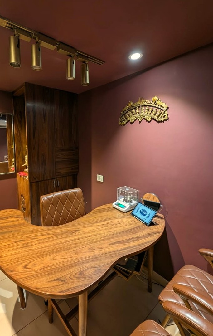 The teak viewing table with the jeweller’s scale in its glass case, a tablet showing a piece, and framed certificates on the wall